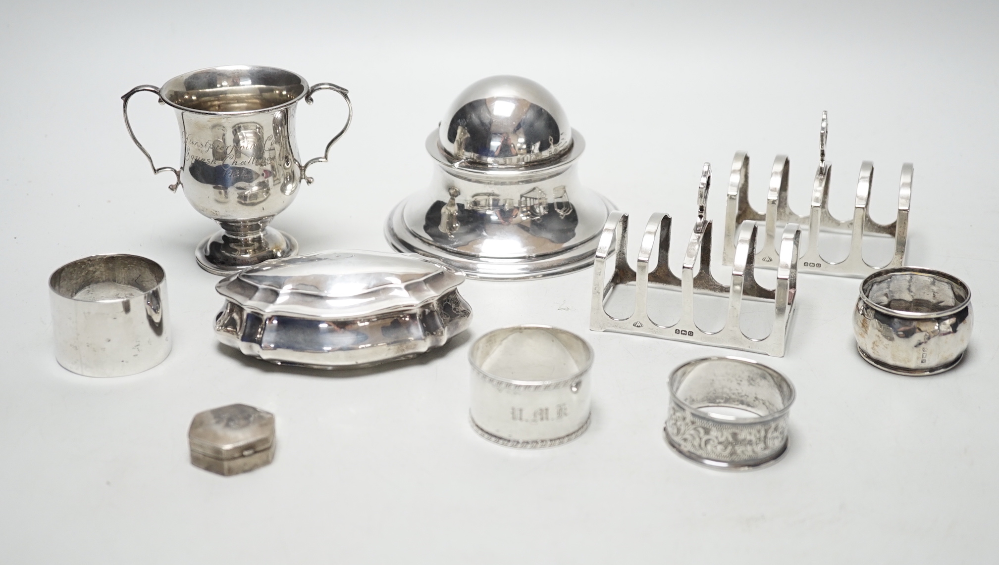 A late Victorian shaped oval silver trinket box, Birmingham, 1898, 99mm, a hexagonal silver pill box, a pair of silver toast racks and four silver serviette rings, a silver domed topped inkwell and a miniature silver two